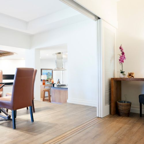 The image shows a modern living area with a dining table, chairs, a desk with a lamp, and a potted plant on a wooden floor.