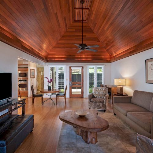 The image shows a living room with wooden ceiling, a TV, couches, a unique coffee table, and artwork on the walls.