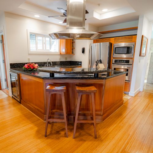 A modern kitchen with wooden flooring, a central island, stainless steel appliances, and two bar stools at the counter.