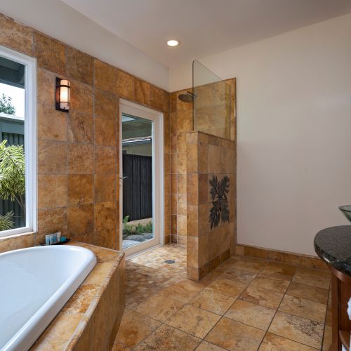 A modern bathroom with a bathtub, glass vessel sink, and walk-in shower, featuring beige tiles and a window view of greenery outside.