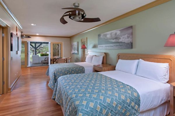 The image shows a hotel room with two double beds, blue patterned bedding, a wood floor, and a seating area. There's a ceiling fan and artwork.