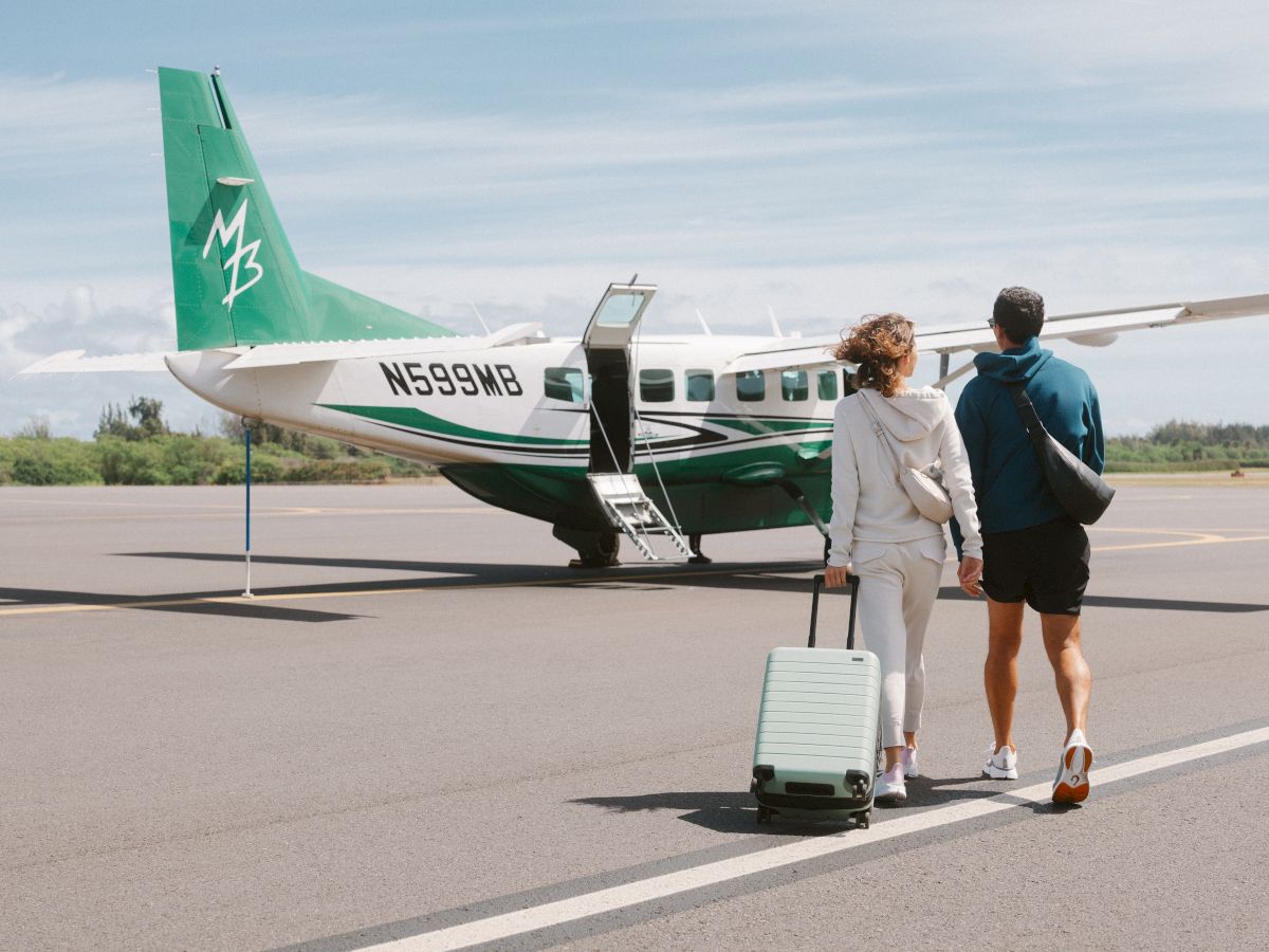 A couple walks towards a small green and white airplane on a runway, with one carrying a suitcase.