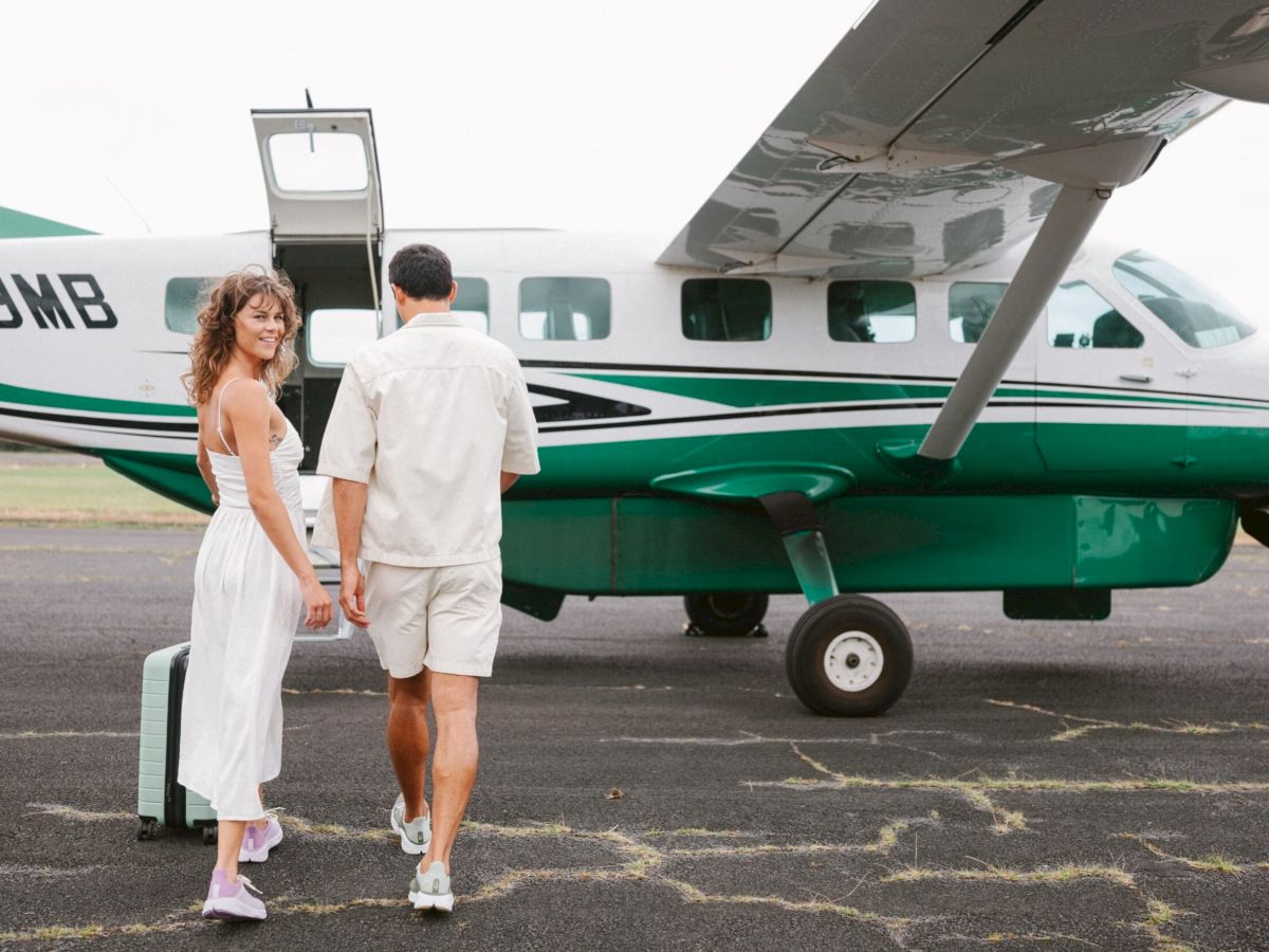 A couple is walking towards a small green and white airplane on an airstrip, with one carrying a suitcase.