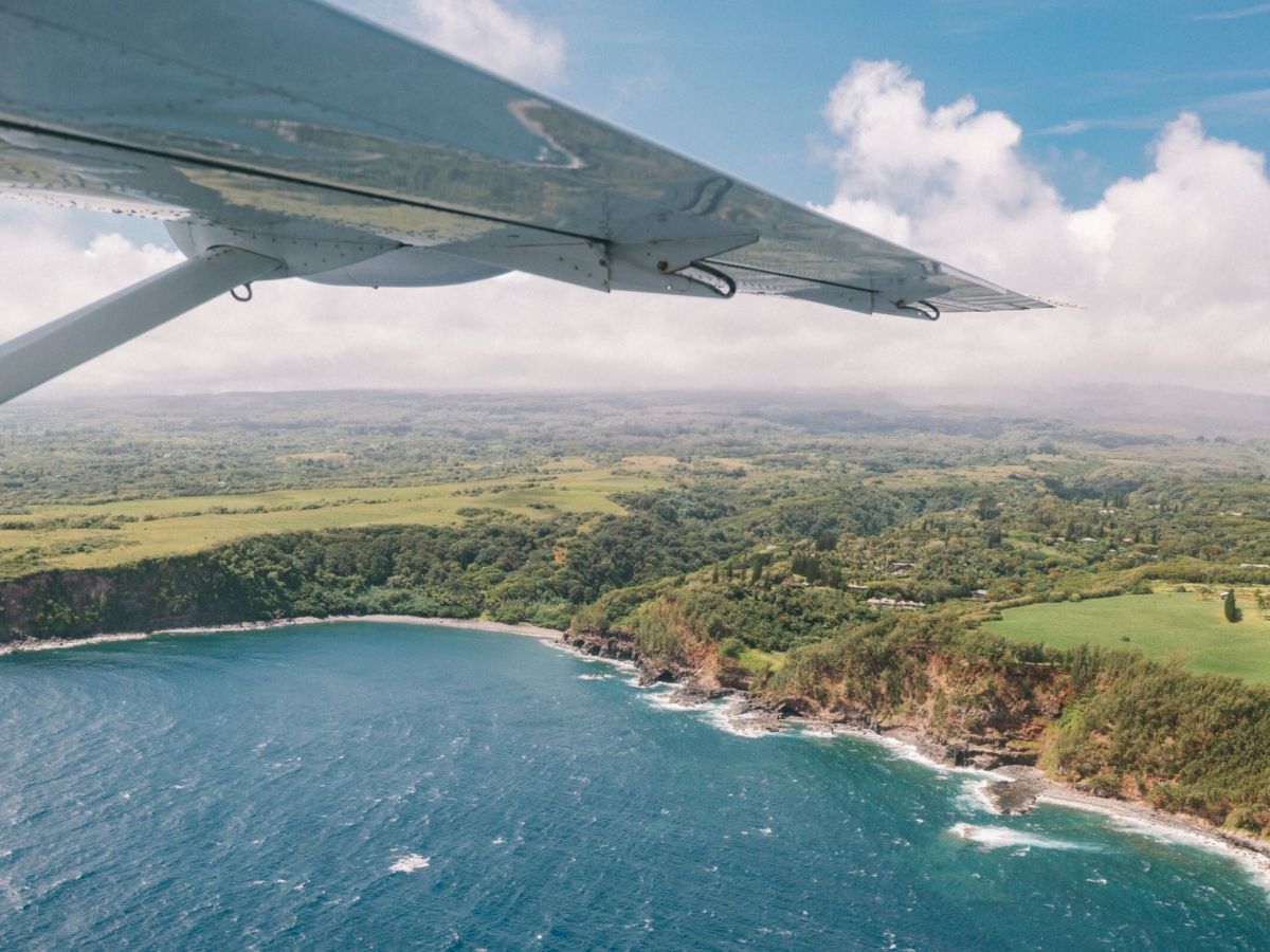 Aerial view of a coastline with lush greenery and ocean waves, seen from a small aircraft wing above.