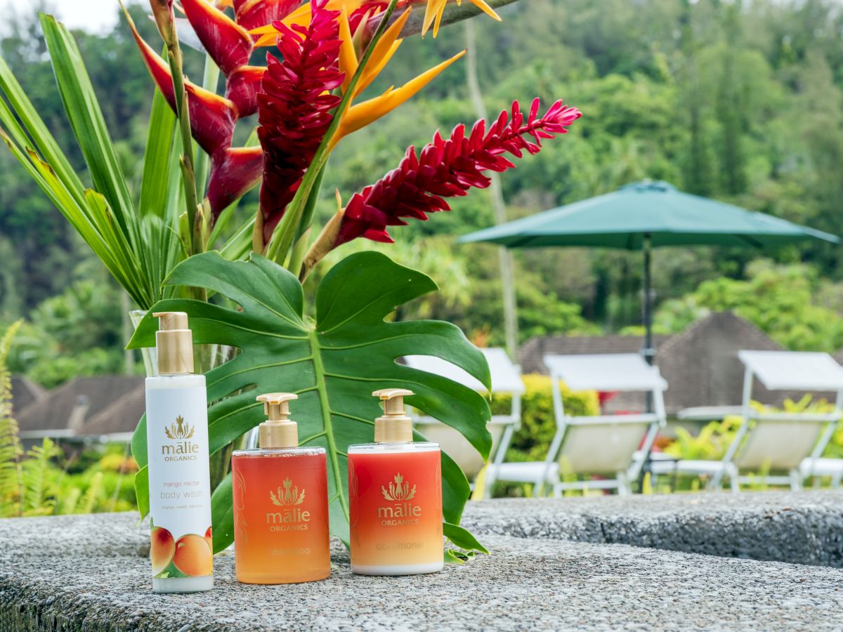 Three Malie Organics products on a stone ledge, with tropical plants and outdoor seating in the background.