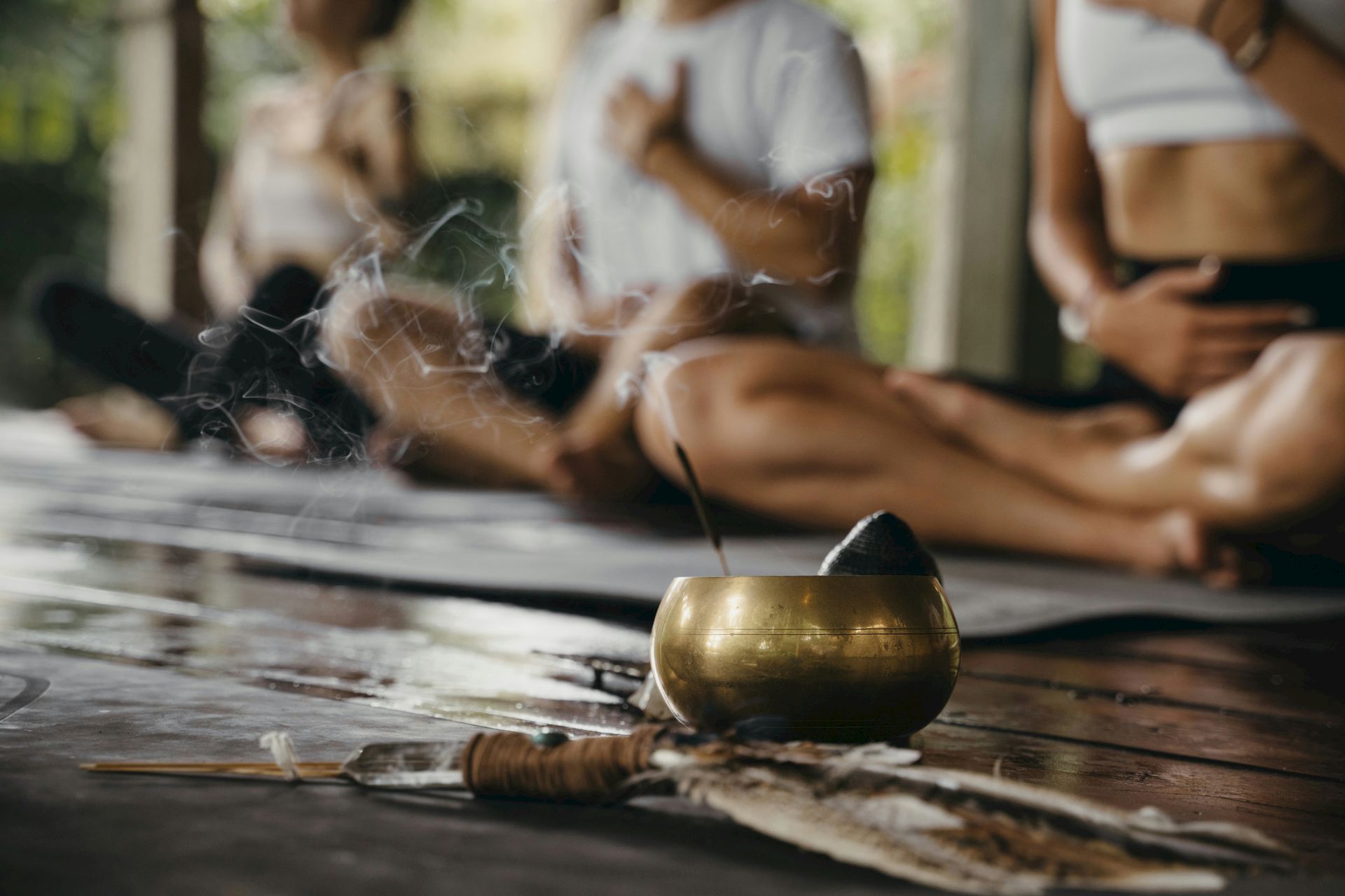 A serene setting with incense burning in a bowl, feathers, and people seated in meditation or yoga poses on mats in the background.