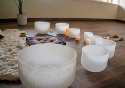 The image shows a set of white crystal singing bowls on a wooden floor, surrounded by candles and a cozy rug, creating a calm ambiance.