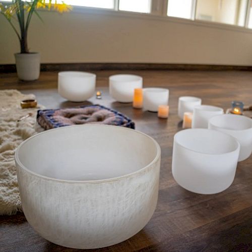 The image shows a set of white crystal singing bowls on a wooden floor, surrounded by candles and a cozy rug, creating a calm ambiance.