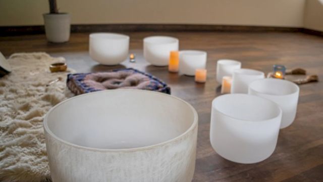 The image shows a set of white crystal singing bowls on a wooden floor, surrounded by candles and a cozy rug, creating a calm ambiance.