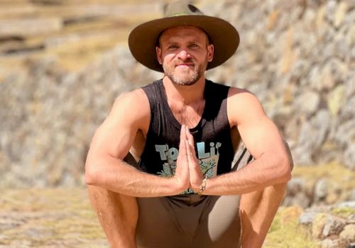 A person is squatting outdoors, wearing a wide-brimmed hat and tank top, with hands pressed together in front, against a stone wall background.