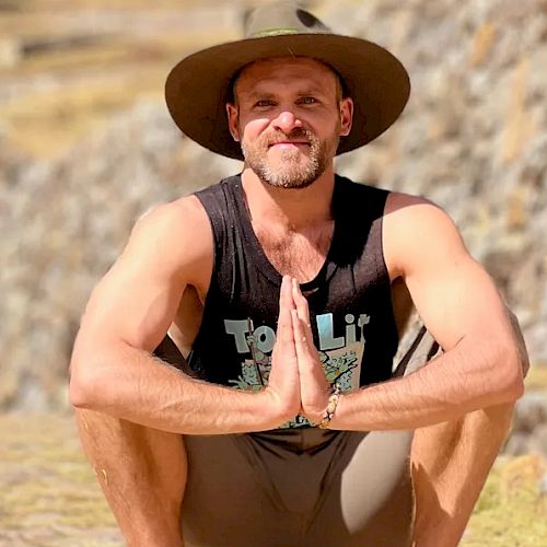 A person is squatting outdoors, wearing a wide-brimmed hat and tank top, with hands pressed together in front, against a stone wall background.