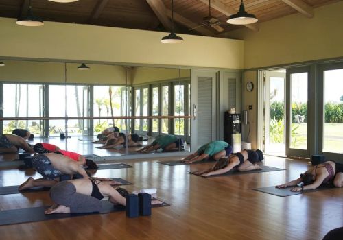 People are practicing yoga in a studio with large windows and mirrors, performing a resting pose on mats on a wooden floor.