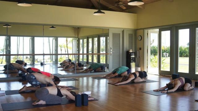 People are practicing yoga in a studio with large windows and mirrors, performing a resting pose on mats on a wooden floor.