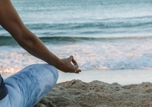 A person seated on the sand in a meditative pose by the ocean, wearing blue pants, with waves in the background.