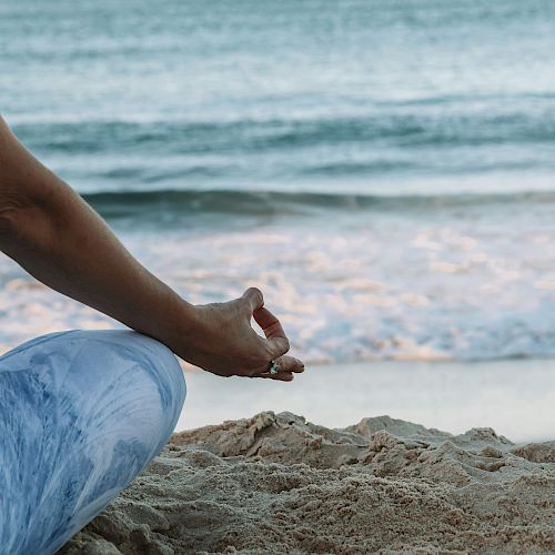 A person seated on the sand in a meditative pose by the ocean, wearing blue pants, with waves in the background.