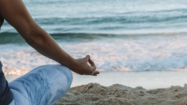 A person seated on the sand in a meditative pose by the ocean, wearing blue pants, with waves in the background.