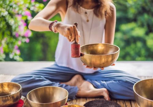 A person is sitting cross-legged, holding a mallet and striking a singing bowl, surrounded by more bowls, in a serene outdoor setting.