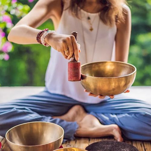 A person is sitting cross-legged, holding a mallet and striking a singing bowl, surrounded by more bowls, in a serene outdoor setting.