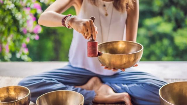 A person is sitting cross-legged, holding a mallet and striking a singing bowl, surrounded by more bowls, in a serene outdoor setting.