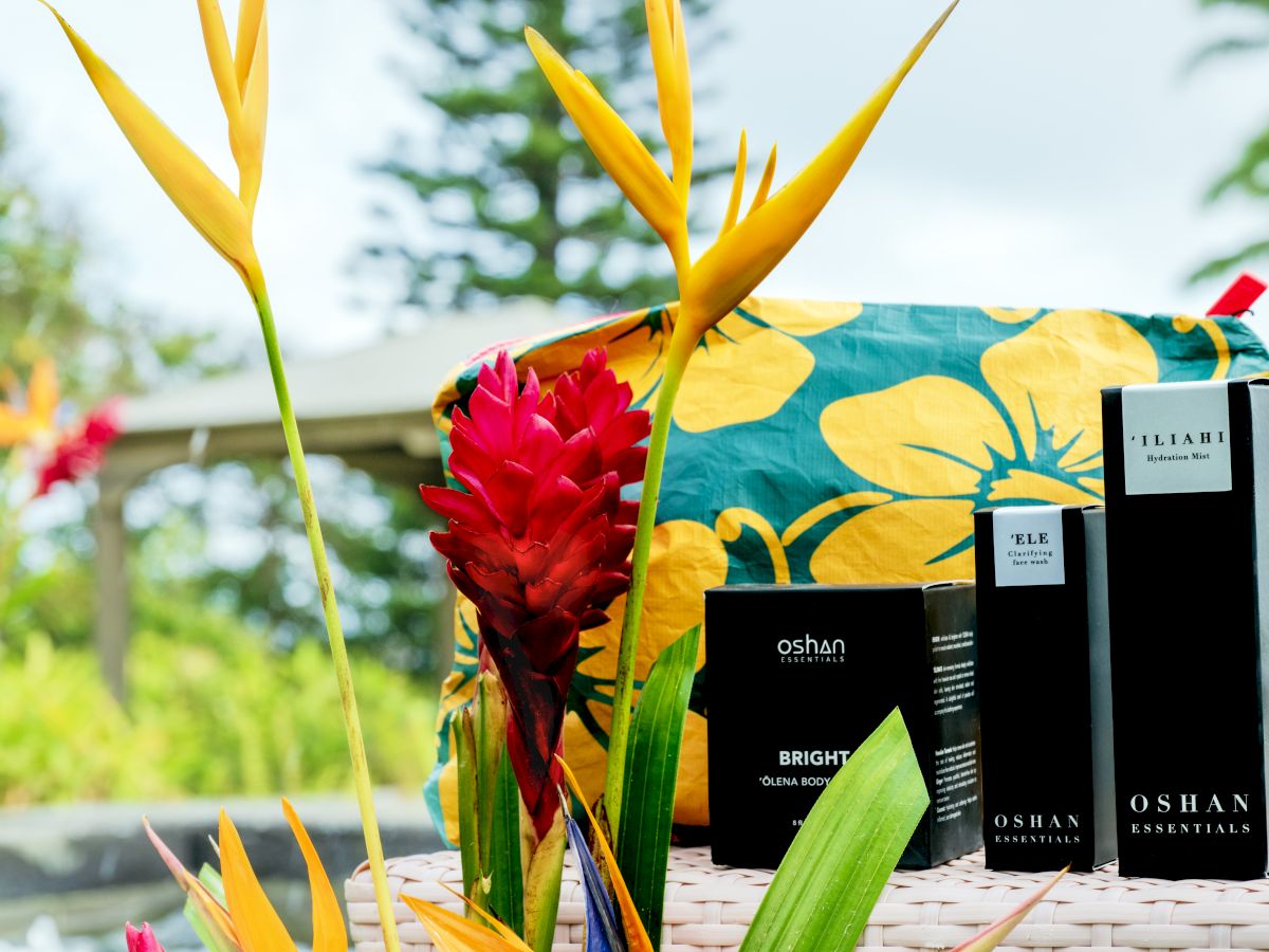 Gift set with skincare products labeled "Oshan Essentials," surrounded by vibrant tropical flowers and a colorful fabric wrap.