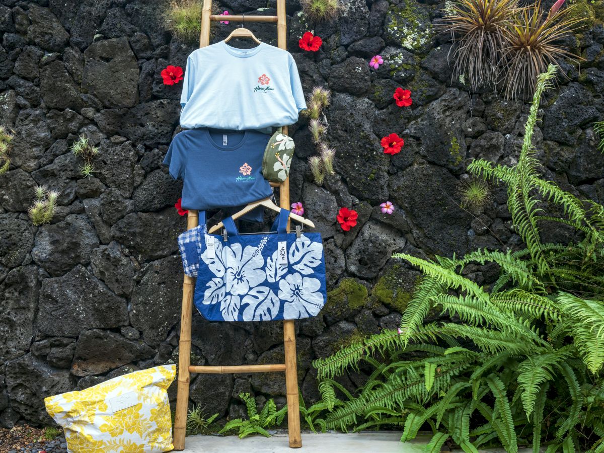 A bamboo ladder displays two shirts and a floral bag against a rocky wall with vibrant flowers and ferns around it.