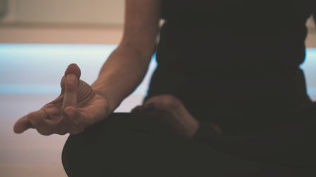 A person is sitting cross-legged in a meditative yoga pose, with one hand resting on the knee and fingers forming a mudra.