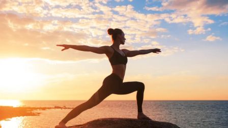 A person is doing yoga in a warrior pose on a rock by the sea during a sunset, with a colorful sky above the water.