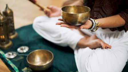 A person is holding a singing bowl and mallet while sitting cross-legged on a mat with other meditation items nearby.