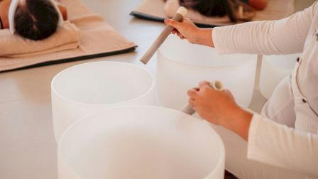 A person plays crystal singing bowls in a room where others are lying on mats, likely participating in a sound therapy session.