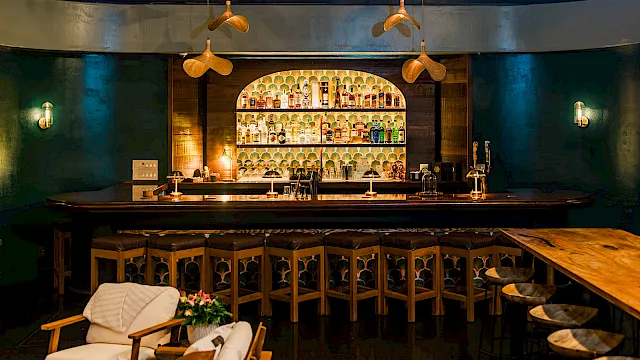 A dimly lit bar with a backlit liquor display, wooden stools, and hanging pendant lights.