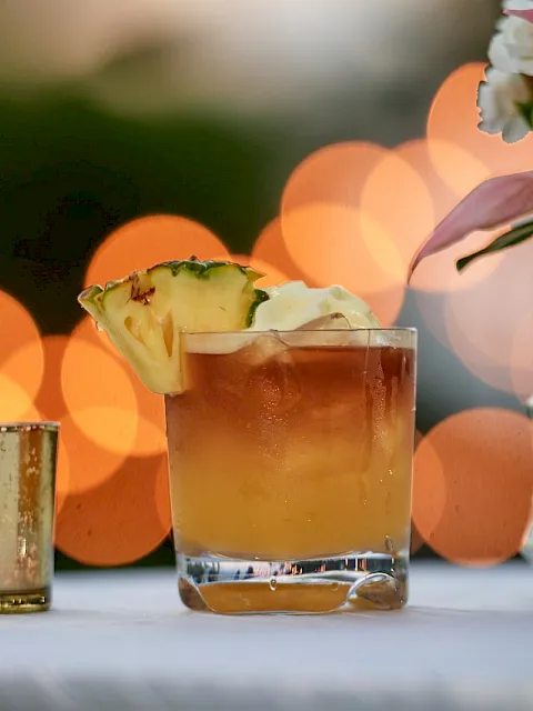 A sunny cocktail with a pineapple garnish sits by a vase of flowers, with warm bokeh lights in the background.