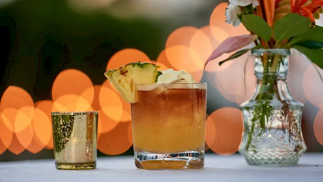 A sunny cocktail with a pineapple garnish sits by a vase of flowers, with warm bokeh lights in the background.