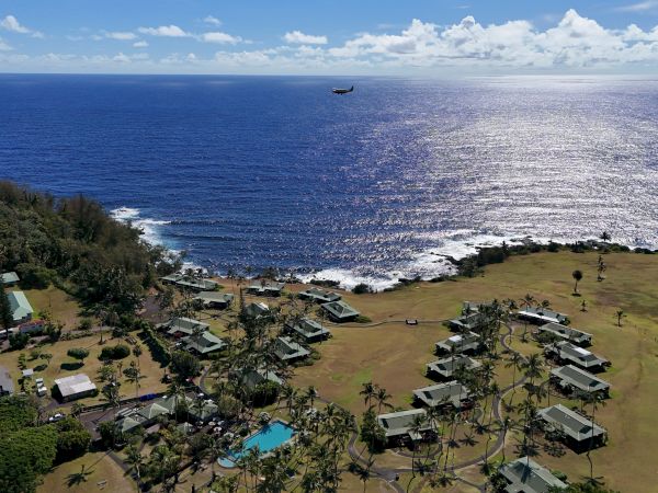 A coastal village with green fields, buildings, and palm trees. The ocean glistens under the sun, with a plane flying above.