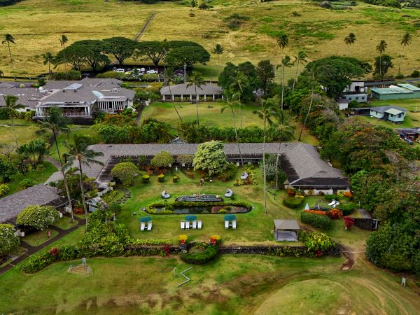Aerial view of a lush landscape featuring several buildings, gardens, and open green spaces surrounded by trees and fields.