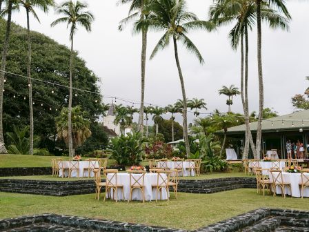 An outdoor venue with round tables and chairs set up on grass, surrounded by palm trees and decorative string lights.