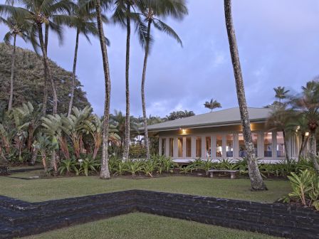A one-story house surrounded by palm trees and lush greenery with a clear sky in the background and a well-kept lawn in the foreground.