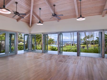 The image shows a spacious room with wooden floors, ceiling fans, and large windows offering a view of palm trees and the ocean.