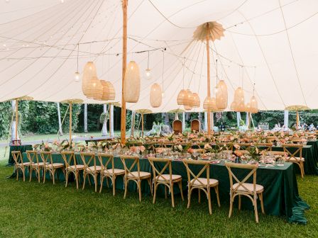 The image shows an elegantly decorated outdoor event tent with long tables, wooden chairs, and pendant lights, set up for a gathering.