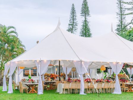 A white tent is set up outdoors with tables and chairs, decorated with floral arrangements and lights, creating an elegant event setting.