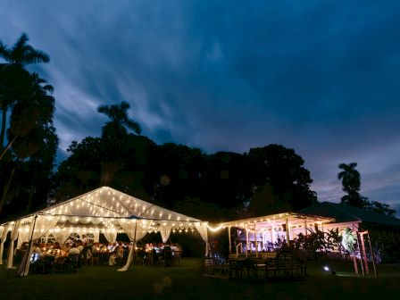 An outdoor event under a lit tent with guests, surrounded by trees and evening sky, creates a festive atmosphere.