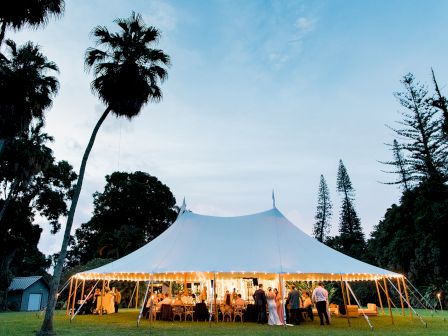 A large, white, well-lit event tent is set up outdoors surrounded by trees, with people gathered inside and near the entrance.