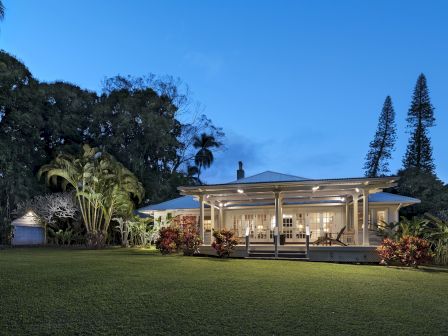 A cozy house with a lit porch is surrounded by greenery, under a clear evening sky with tall trees in the background.