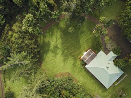 An aerial view of a house with a green roof, surrounded by lush trees and a pathway on a large grassy area.