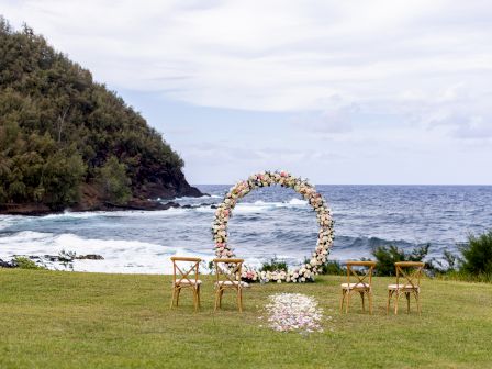 A scenic seaside setup with a floral arch and four chairs on a lawn, perfect for a small outdoor ceremony by the ocean.