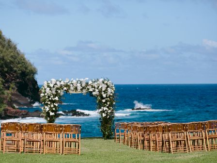 An outdoor wedding setup by the ocean, featuring a flower-adorned arch and rows of wooden chairs on green grass.