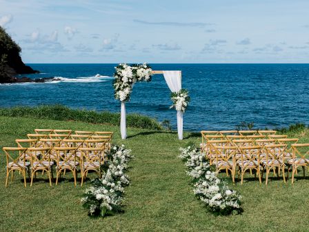 An outdoor wedding setup features wooden chairs and a flower-decorated arch by the ocean, with greenery and blue skies in the background.