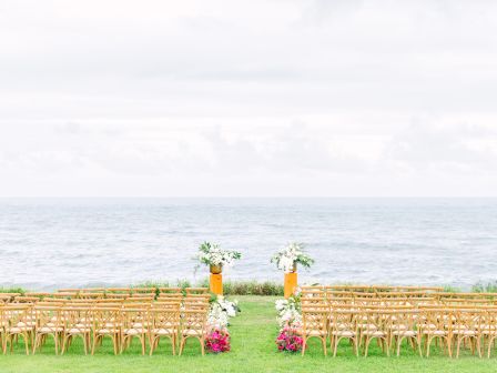 Outdoor wedding setup with rows of chairs facing an ocean view, decorated archway, and flower arrangements on a grassy area.