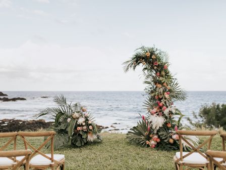 An oceanside wedding setup with wooden chairs, floral arch, and greenery on a grassy area overlooking the sea under a cloudy sky.