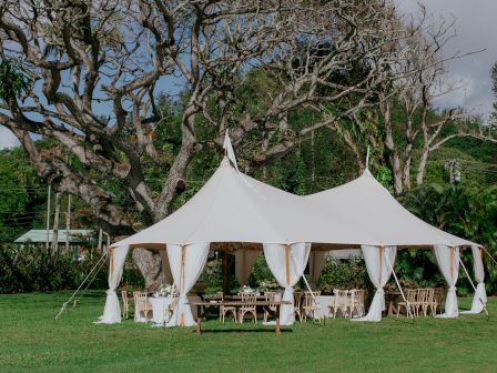 A large white event tent with open sides is set up on grass, surrounded by trees, with chairs and tables arranged inside for a gathering.