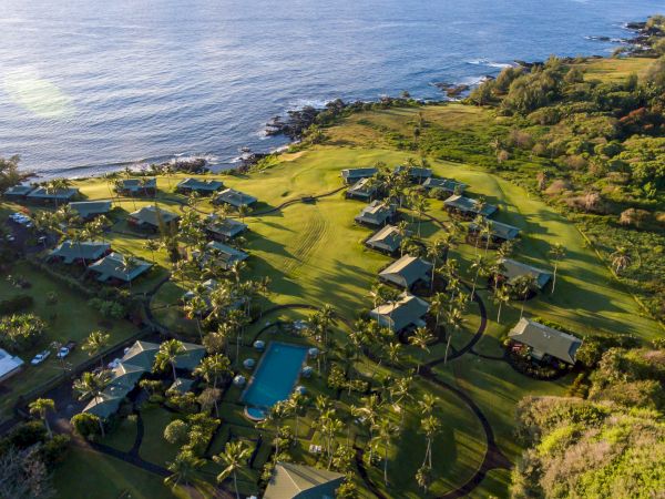 An aerial view of a tropical beachfront resort with villas and palms along a winding coast beside the blue ocean.